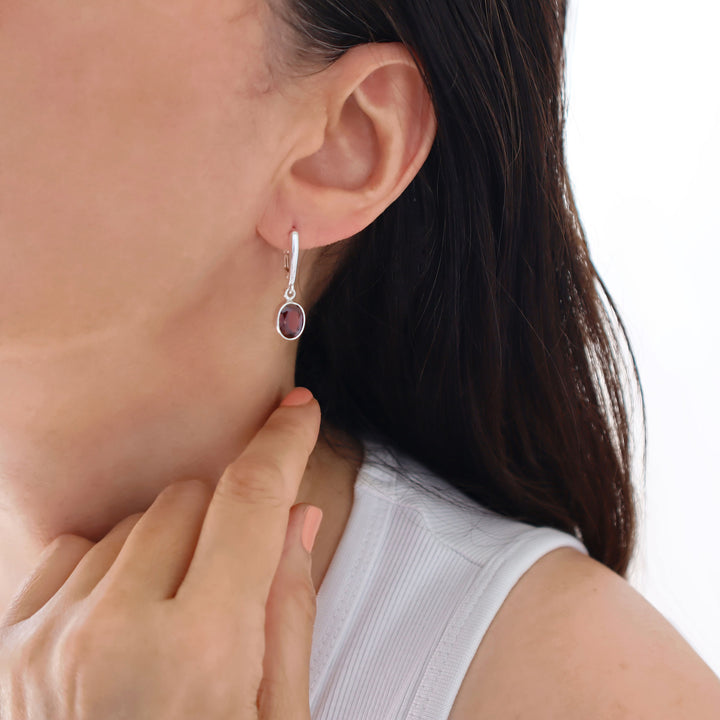 A close-up of a female model wearing a silver dangling drop earring with a small oval dark red faceted garnet gemstone.