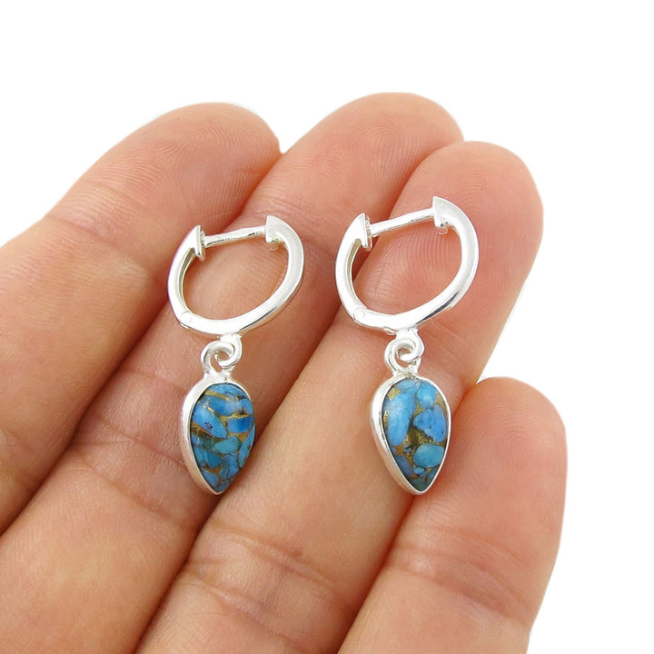 A pair of 925 silver huggie hoop earrings with teardrop-shaped blue turquoise stone drops, shown resting on a person's fingers against a white background.