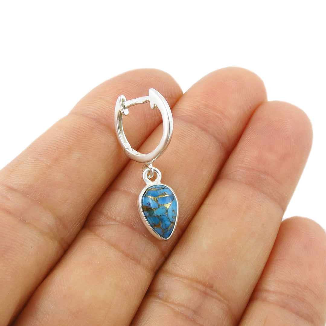 A sterling silver huggie hoop earring with a dangling teardrop-shaped blue turquoise stone, held in the centre of a person's hand against a white background.