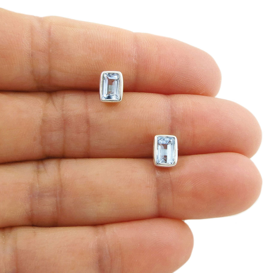 A pair of silver stud earrings with rectangular blue topaz gemstones, shown resting on a person's fingers against a white background.