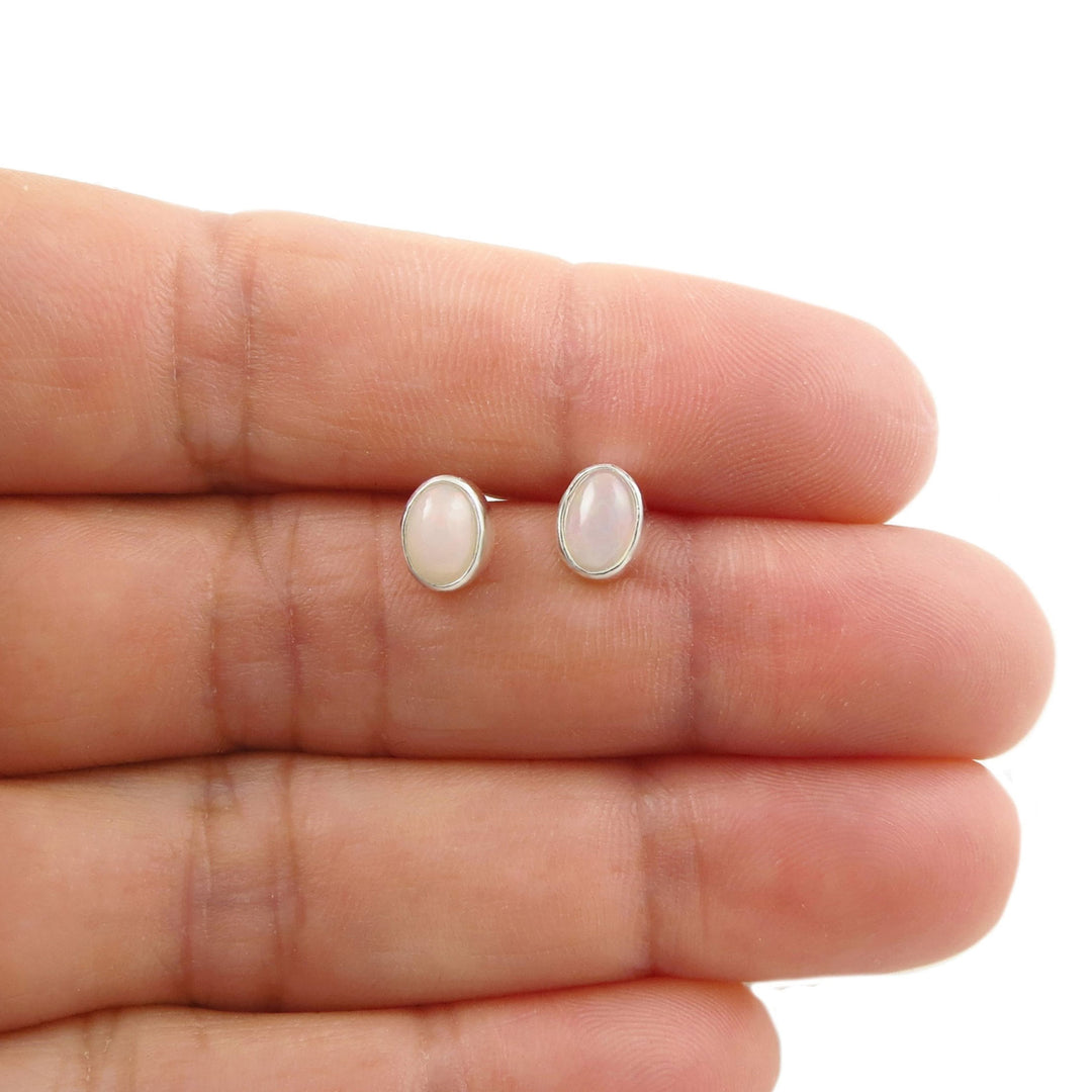 A pair of oval stud earrings with milky white Ethiopian opal cabochon stones in silver settings, shown resting on a person's fingers against a white background.
