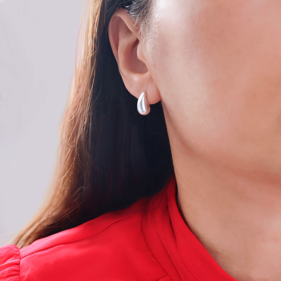 A close-up of a woman's ear wearing a small, polished silver teardrop-shaped stud earring, with dark hair and a bright red top visible against a light gray background.