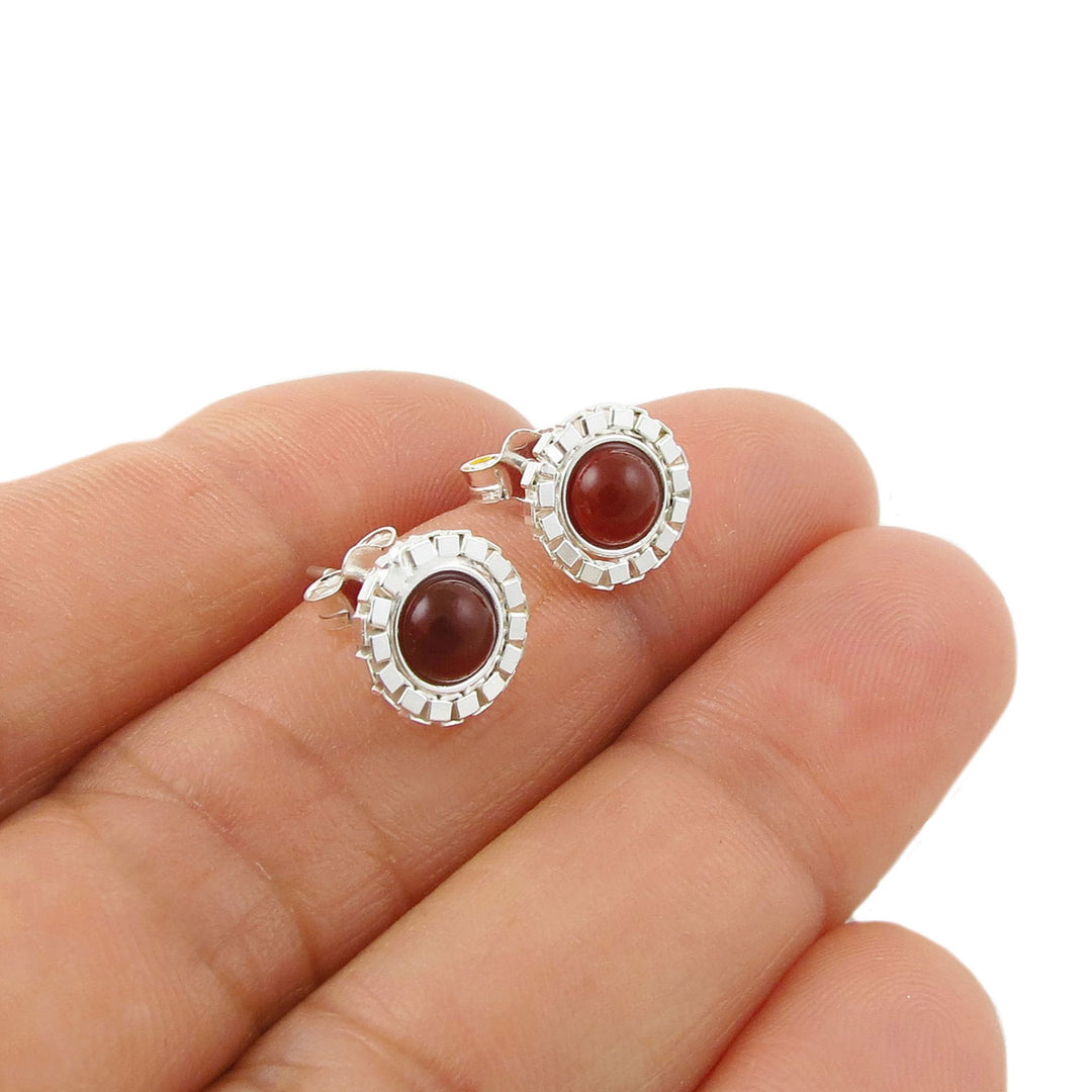 A pair of silver stud earrings with oval reddish-brown carnelian cabochon gemstones and textured silver borders, shown resting on a person's fingers against a white background.