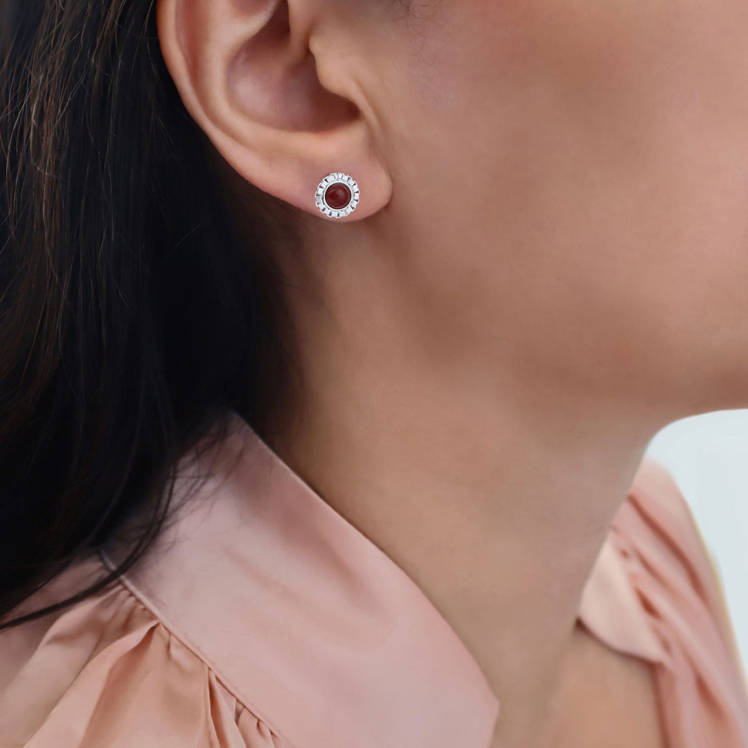 A close-up of a model's ear wearing a small circular stud 925 silver earring with a deep red carnelian gemstone centre stone.