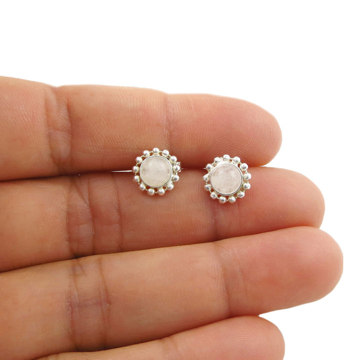 A pair of silver stud earrings with round pale rock crystal quartz stones and a beaded halo border, shown resting on a person's fingers against a white background.