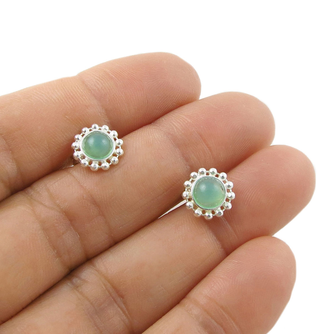 A pair of 925 silver stud earrings with green calcedony chrysoprase gemstones and a beaded halo border, shown resting on a person's fingers against a white background.