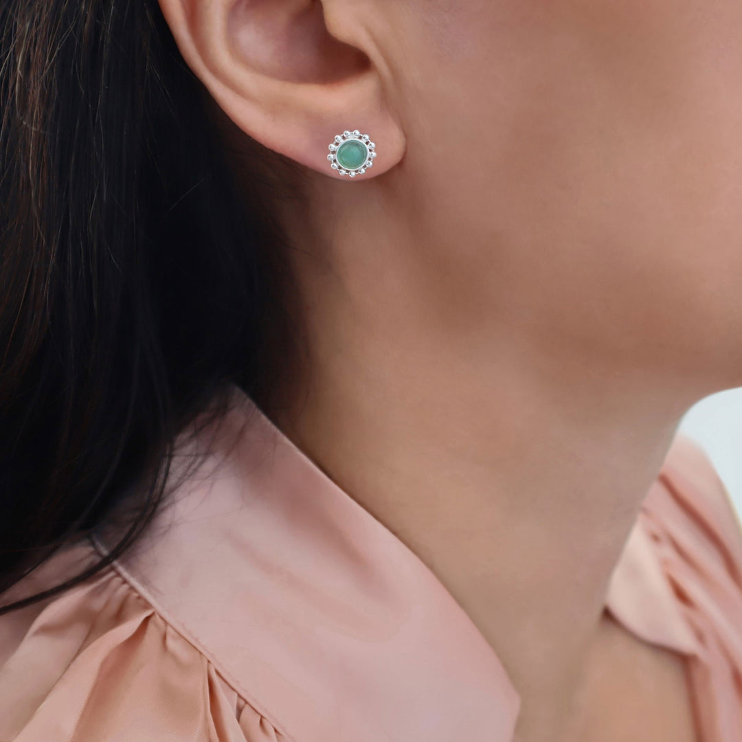 A close-up of a woman's ear wearing a small round stud earring with a green chrysoprase gemstone centre and a halo of clear stones, paired with a light pink blouse.