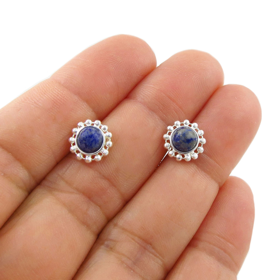 A pair of 925 silver stud earrings with round blue lapis lazuli stones and a beaded halo border, shown resting on a person's palm against a white background.
