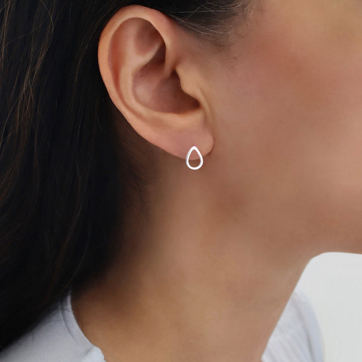 A close-up of a woman's ear wearing a small, minimalist teardrop-shaped stud earring in bright 925 silver.
