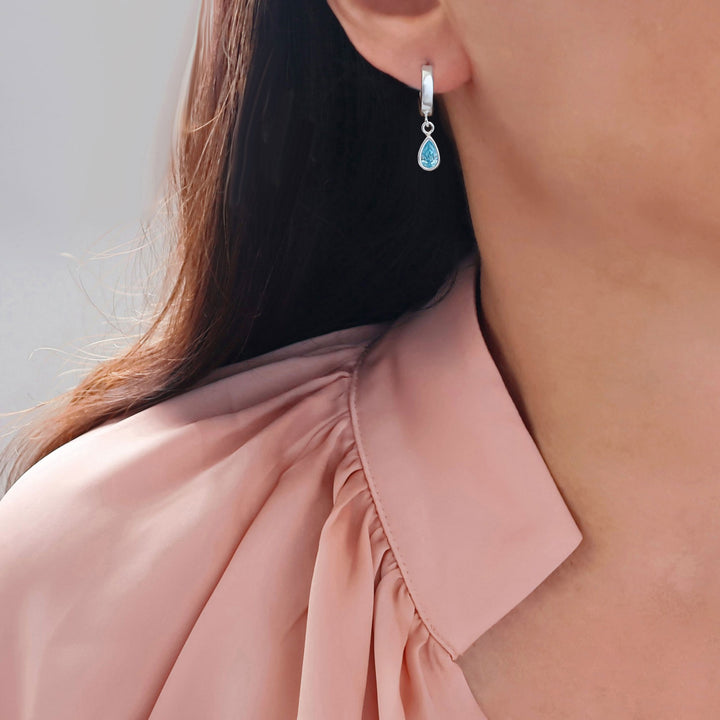 A close-up of a woman wearing a sterling 925 silver huggie hoop earring with a teardrop-shaped light blue aquamarine gemstone, paired with a dusty pink blouse.