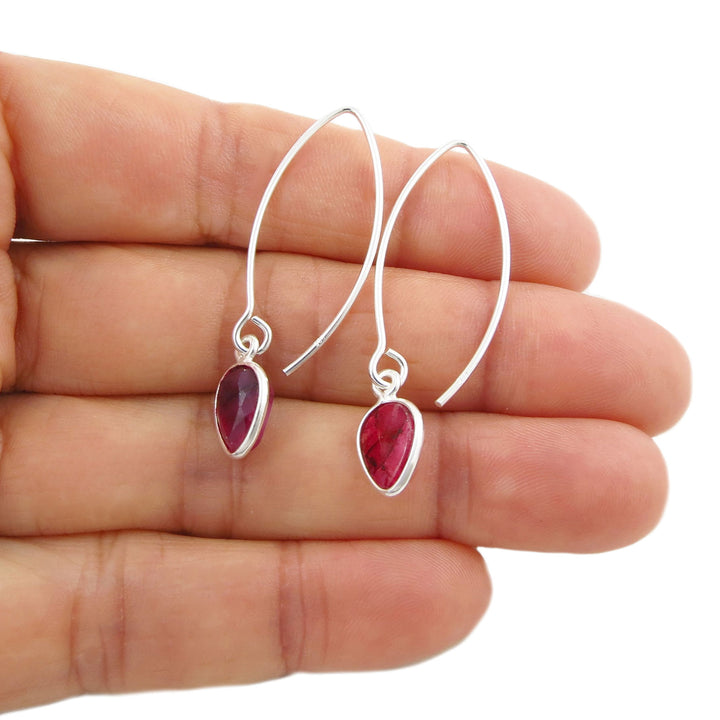 A pair of silver drop earrings with teardrop-shaped pinkish-red faceted ruby quartz gemstones, shown resting on a person's hand against a white background.
