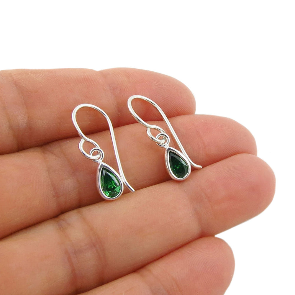 A pair of silver drop earrings with teardrop-shaped green emerald gemstones, held in a person's hand against a plain white background.