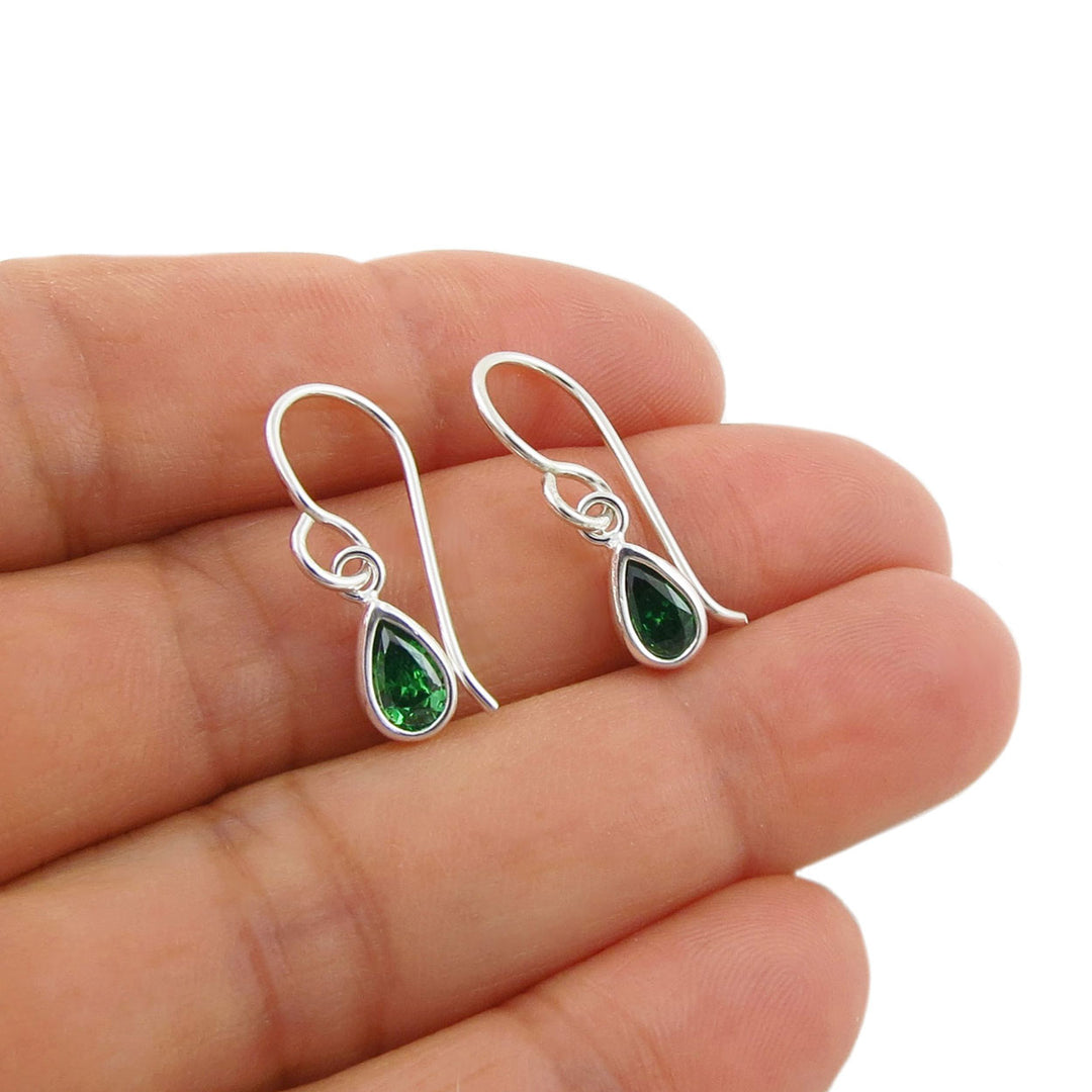 A pair of silver drop earrings with teardrop-shaped green emerald gemstones, held in a person's hand against a plain white background.