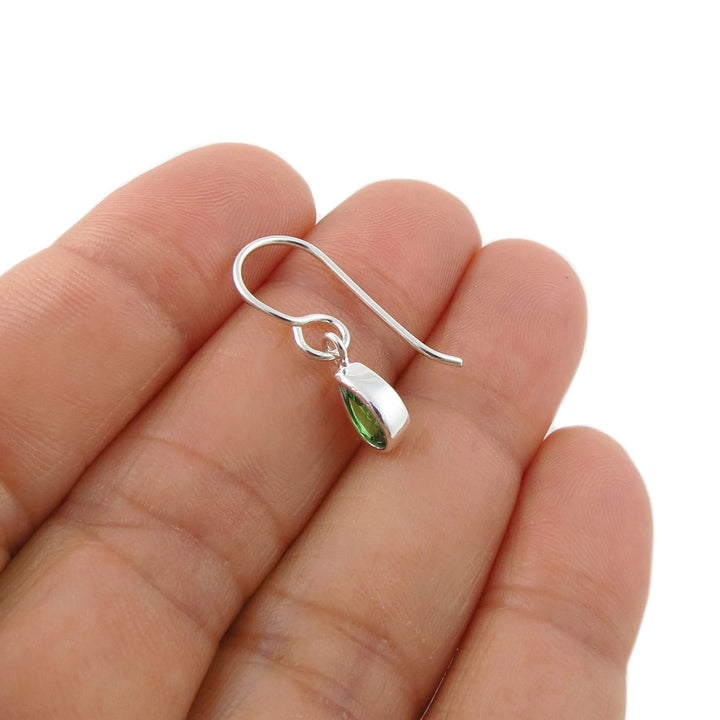A sterling silver fishhook earring with a small marquise-cut green emerald gemstone, held on a person's fingers against a plain white background.