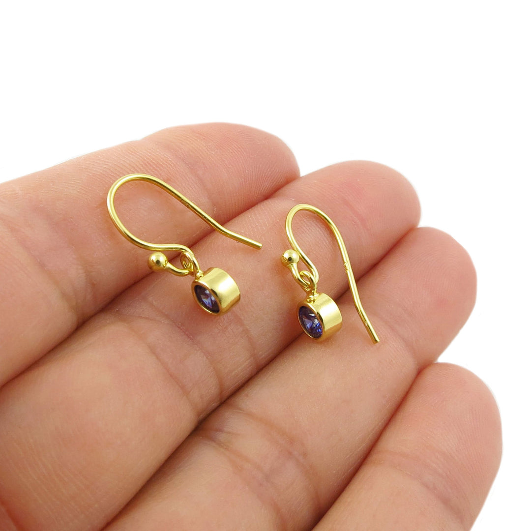 A pair of yellow gold drop earrings with small oval blue sapphire gemstones, shown resting on a person's fingers against a white background.