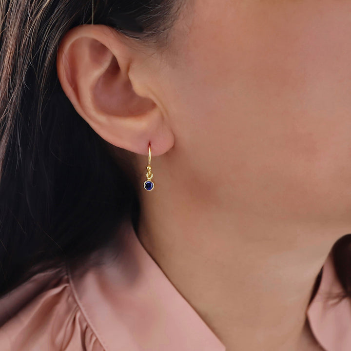 A close-up of a woman's ear wearing a delicate yellow gold dangle earring with a small round dark blue sapphire gemstone, paired with a pink satin blouse.