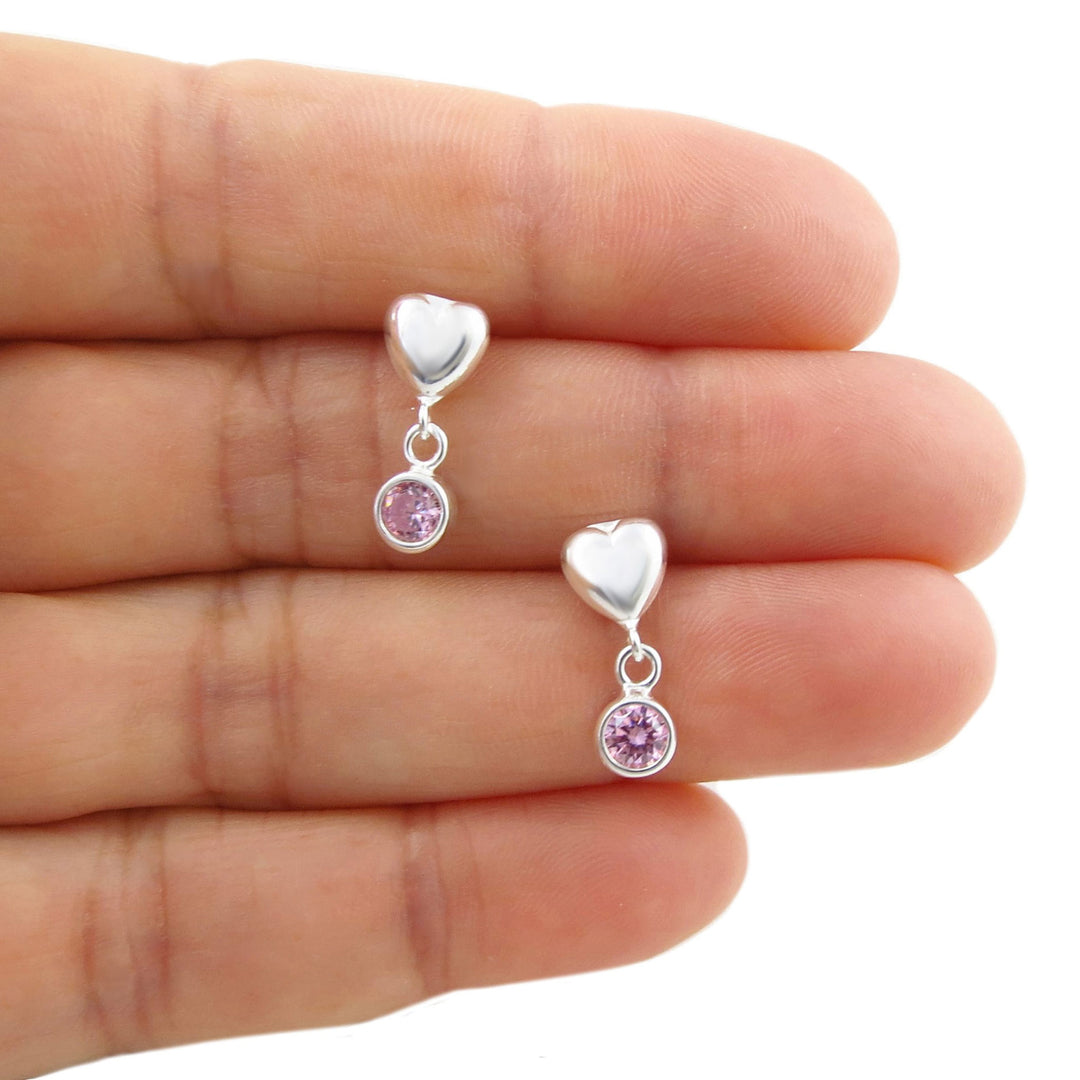 A pair of silver heart dangle earrings with small round pink alexandrite gemstones, shown resting on a person's fingers against a white background.