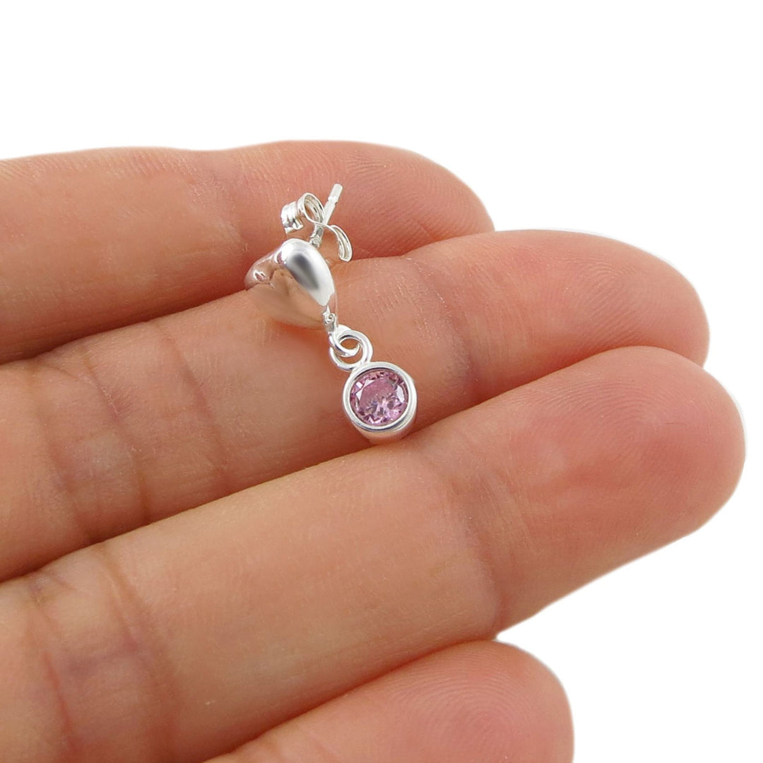 A 925 silver heart-shaped dangle earring with a round pink alexandrite gemstone, shown resting on a person's fingers against a white background.