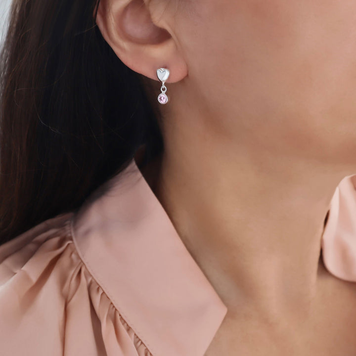 A close-up of a model wearing a heart shaped 925 silver dangle earring with a dangling pink alexandrite gemstone, paired with a light pink blouse.
