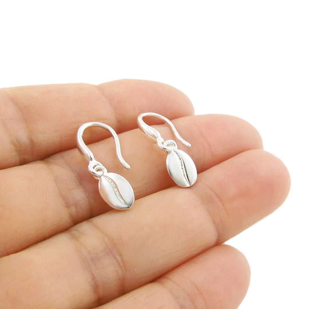 A pair of small silver fish-hook earrings with oval cocoa coffee bean charms, displayed on a person's fingers against a white background.