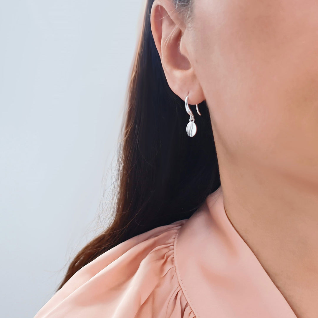 A close-up of a woman's ear wearing a delicate silver dangling coffee bean earring paired with a soft peach-colored blouse against a light gray background.