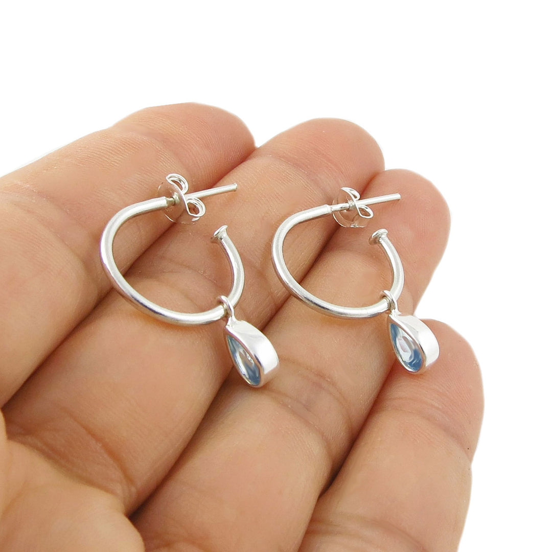 A pair of 925 silver crescent mid-sized hoop earrings with teardrop-shaped light blue aquamarine March birthstone gemstones, shown resting on a woman's fingers against a white background.