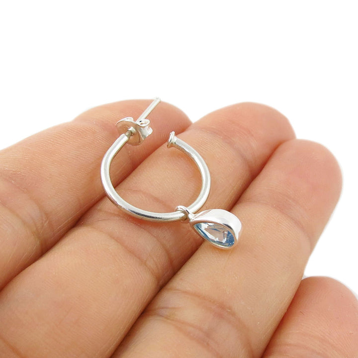 A medium sized 925 silver hoop earring with a teardrop-shaped light blue aquamarine gemstone, shown resting on a woman's fingers against a white background.