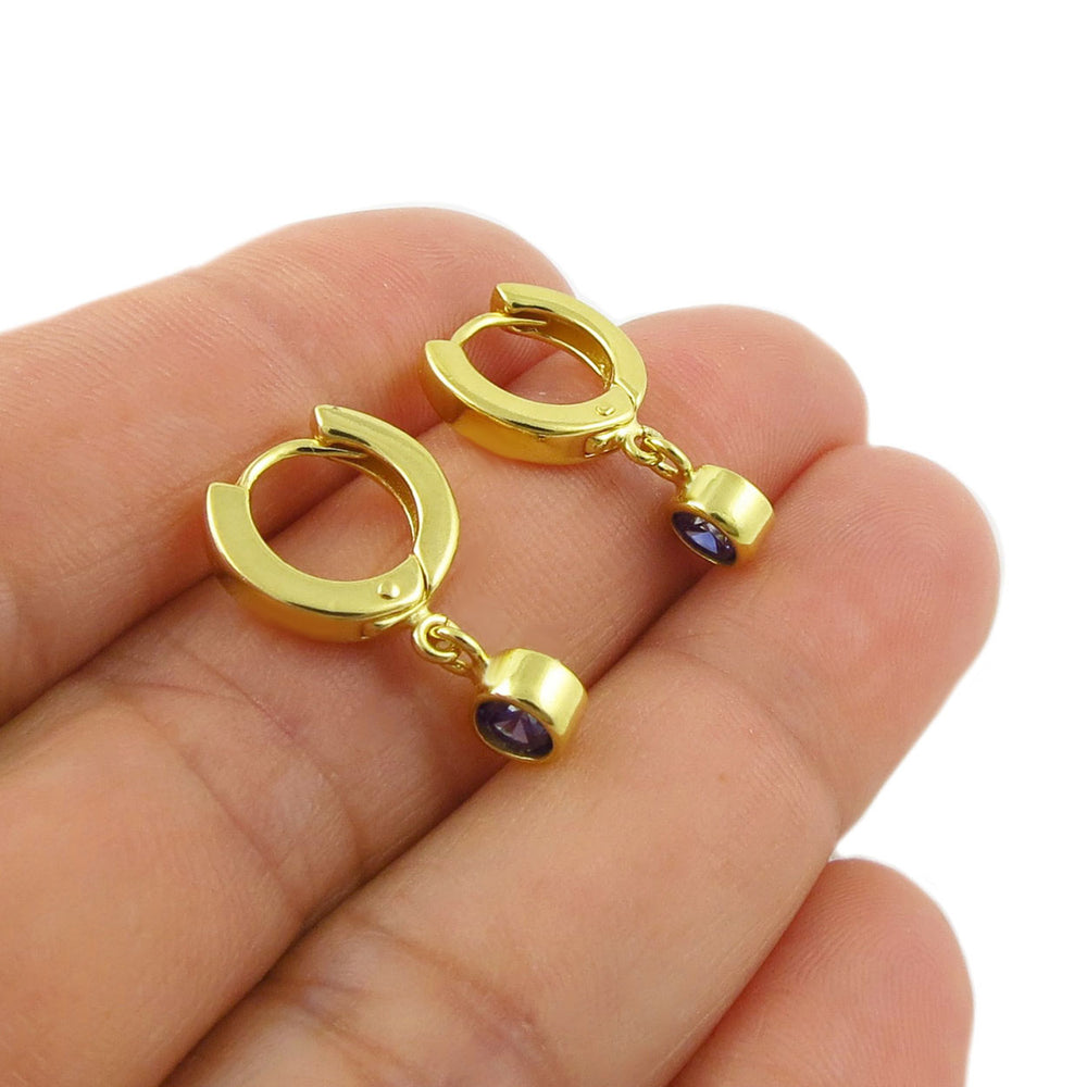 A pair of gold huggie hoop earrings with small blue sapphire gemstone drops, shown resting on a person's fingers against a white background.