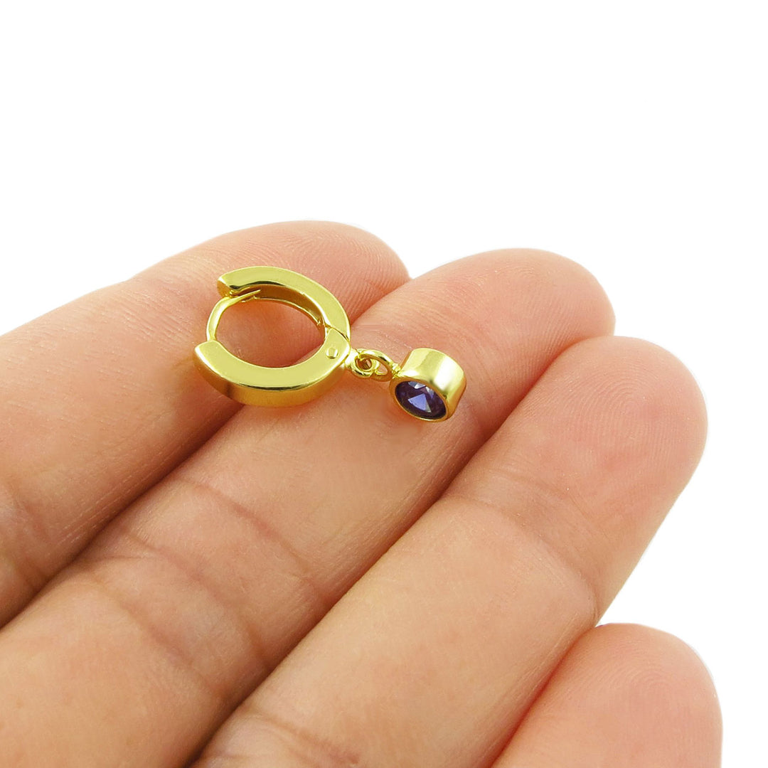 A small gold huggie hoop earring with a dangling teardrop-shaped dark blue sapphire gemstone, shown resting on a person's fingers against a white background.