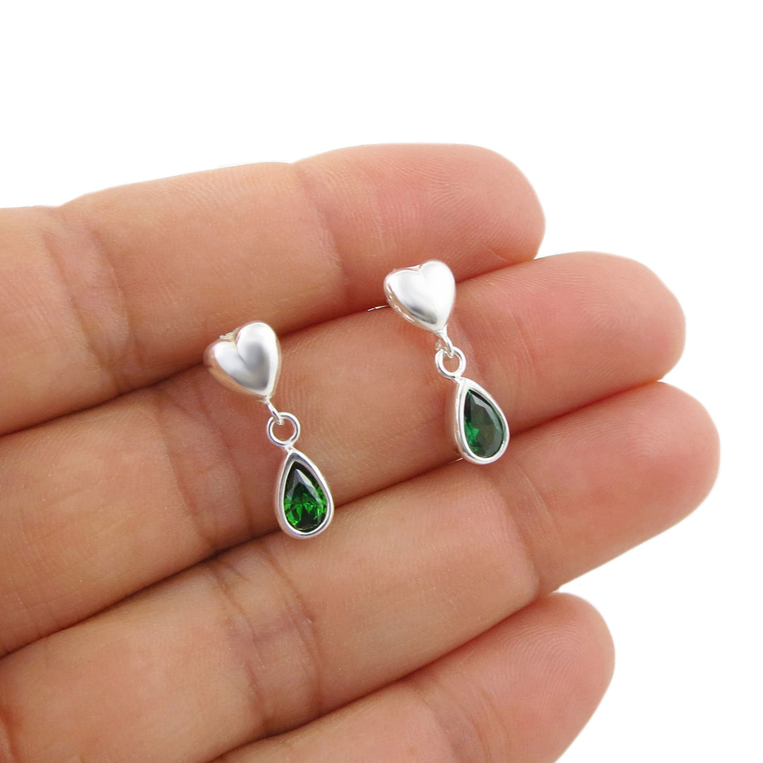 A pair of silver heart and teardrop dangle earrings with green emerald gemstones, shown resting on a person's hand against a white background.