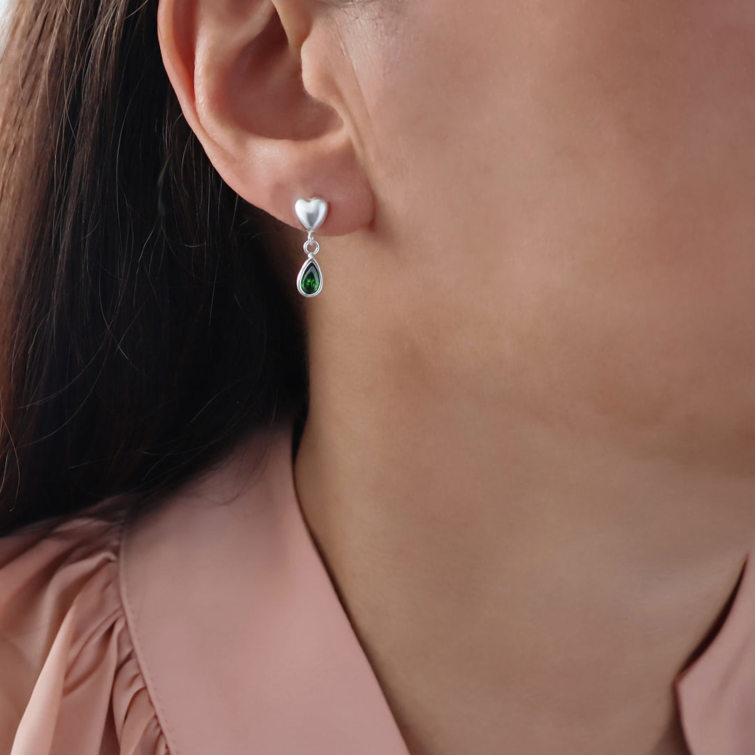 A close-up of a woman's ear wearing a silver drop earring with a small heart-shaped stud and a teardrop green emerald gemstone.