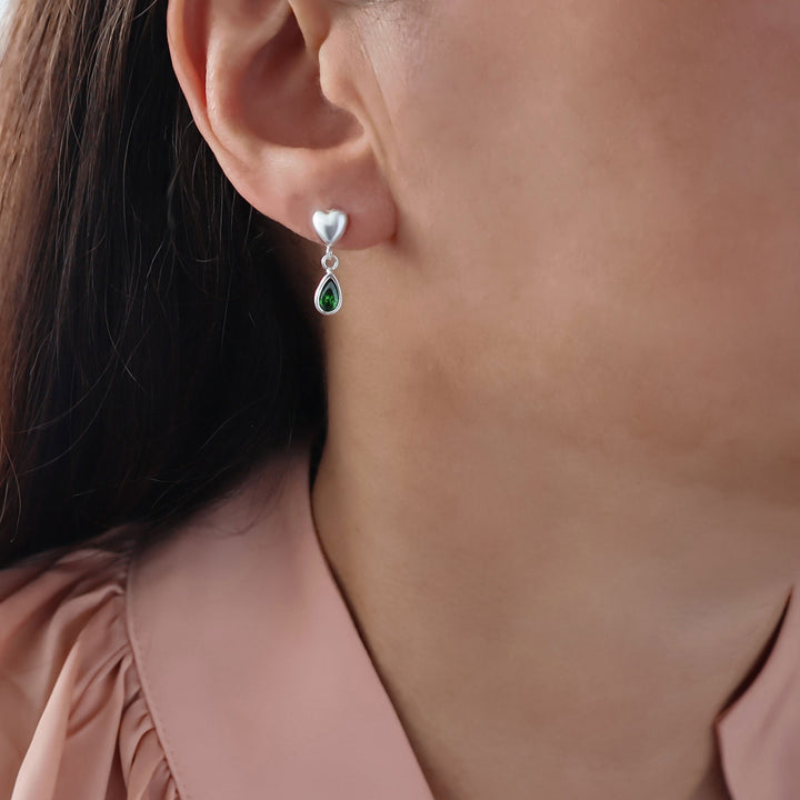 A close-up of a woman's ear wearing a silver drop earring with a small heart-shaped stud and a teardrop green emerald gemstone.