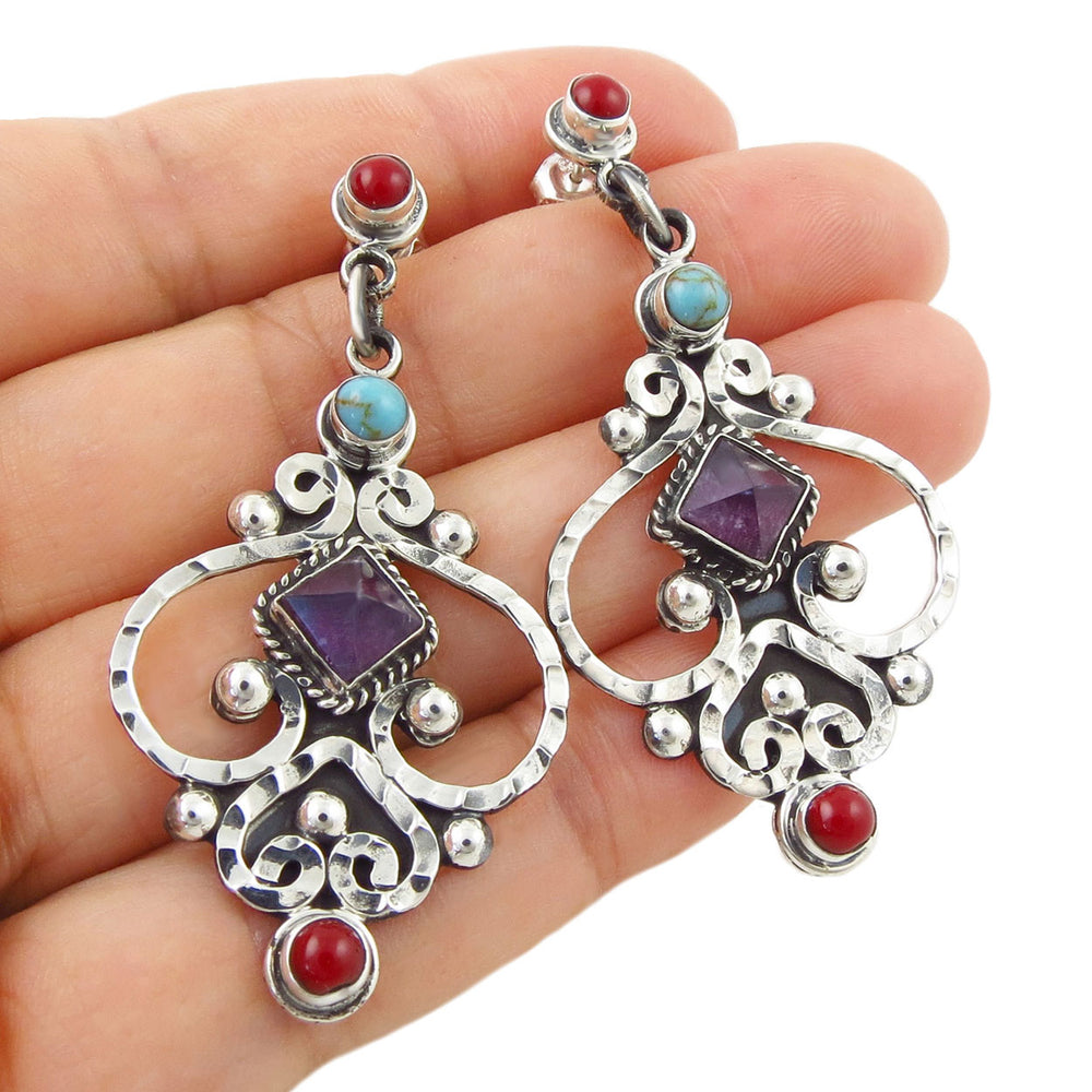 A pair of ornate Mexican 925 sterling silver dangling chandelier style earrings with red, turquoise, and purple amethyst  gemstones, shown resting on a person's hand against a white background.