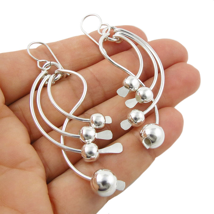A pair of Mexican 925 silver dangling earrings with sculpted sticks decorated with bead accents, shown resting on a person's hand against a white background.