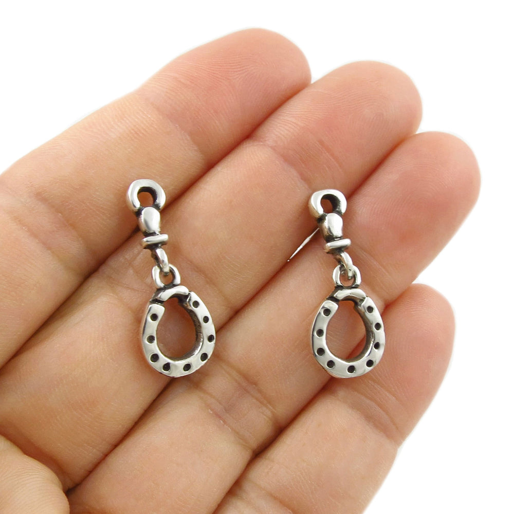 A pair of sterling 925 silver equestrian horseshoe dangle earrings held in a person's hand against a white background.