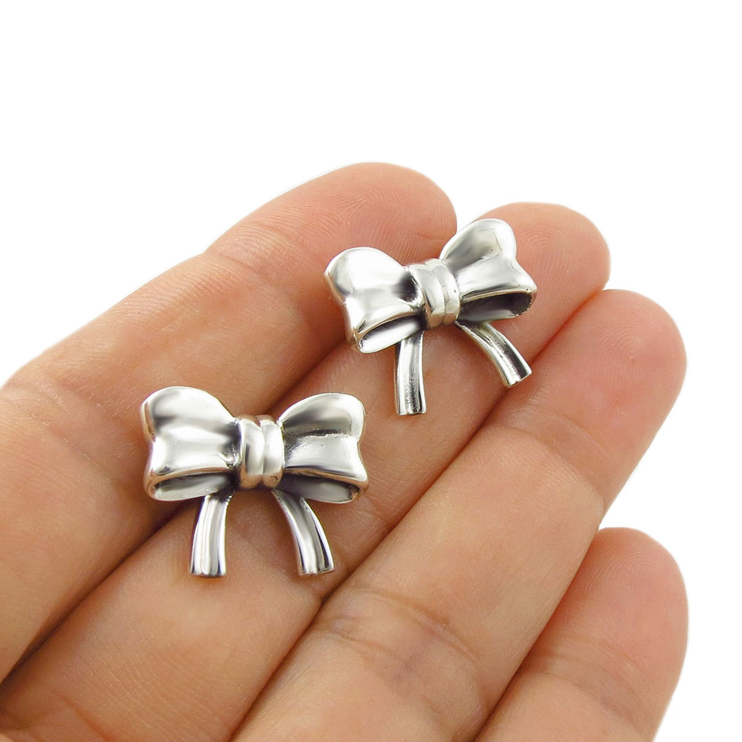 Two mid-sized 925 sterling silver ribbon bow shaped stud earrings resting on a person's fingers against a plain white background.
