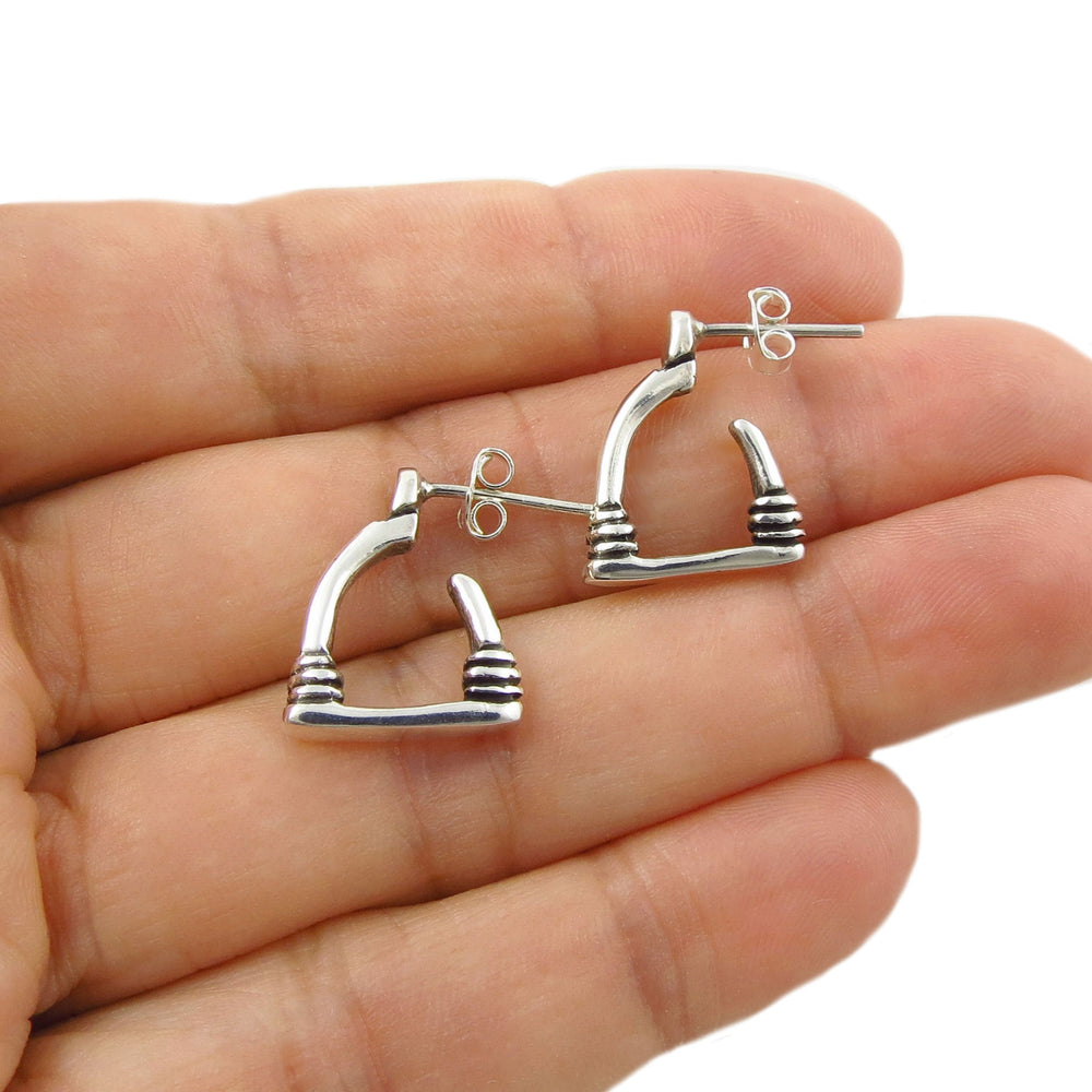 A pair of silver hoop earrings designed like textured equestrian horse stirrups, shown resting on an open hand against a white background.