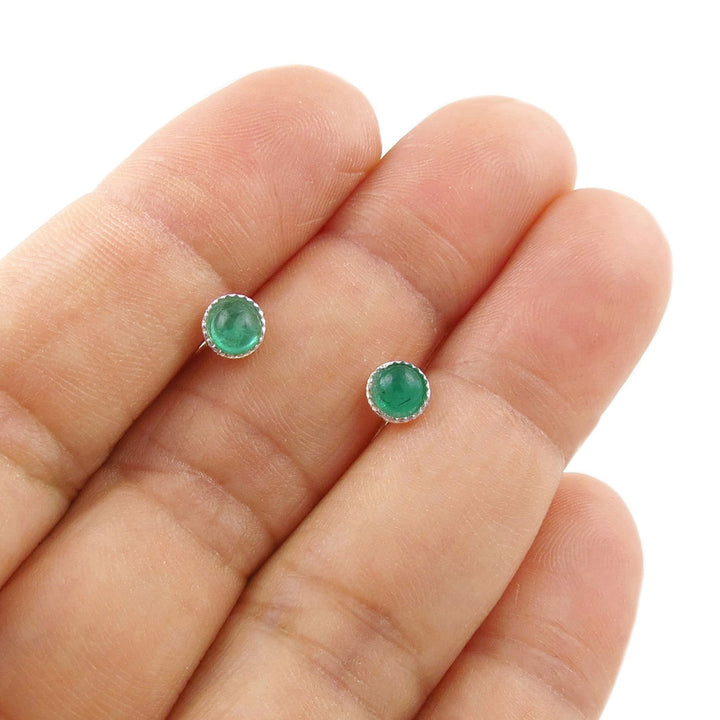 A pair of small round green emerald cabochon stud earrings in silver settings, shown resting on a person's fingers against a white background.