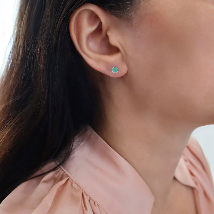 A close-up of a woman's ear wearing a small round stud earring with a vibrant green natural emerald gemstone at the centre, paired with a soft pink blouse.