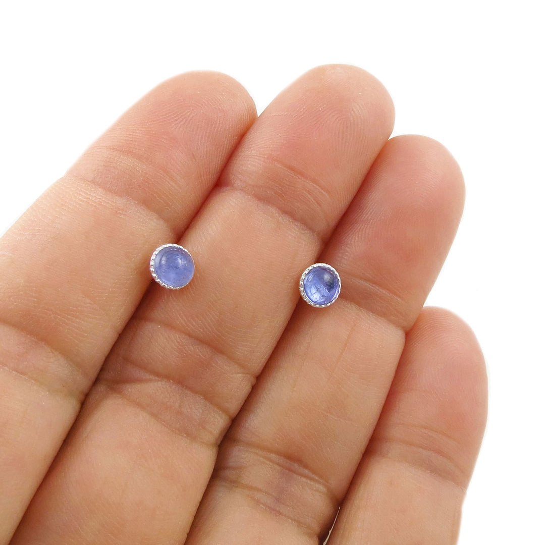 A pair of circle stud earrings with light blue tanzanite cabochon gemstones in silver bezel settings, shown resting on a person's fingers against a white background.