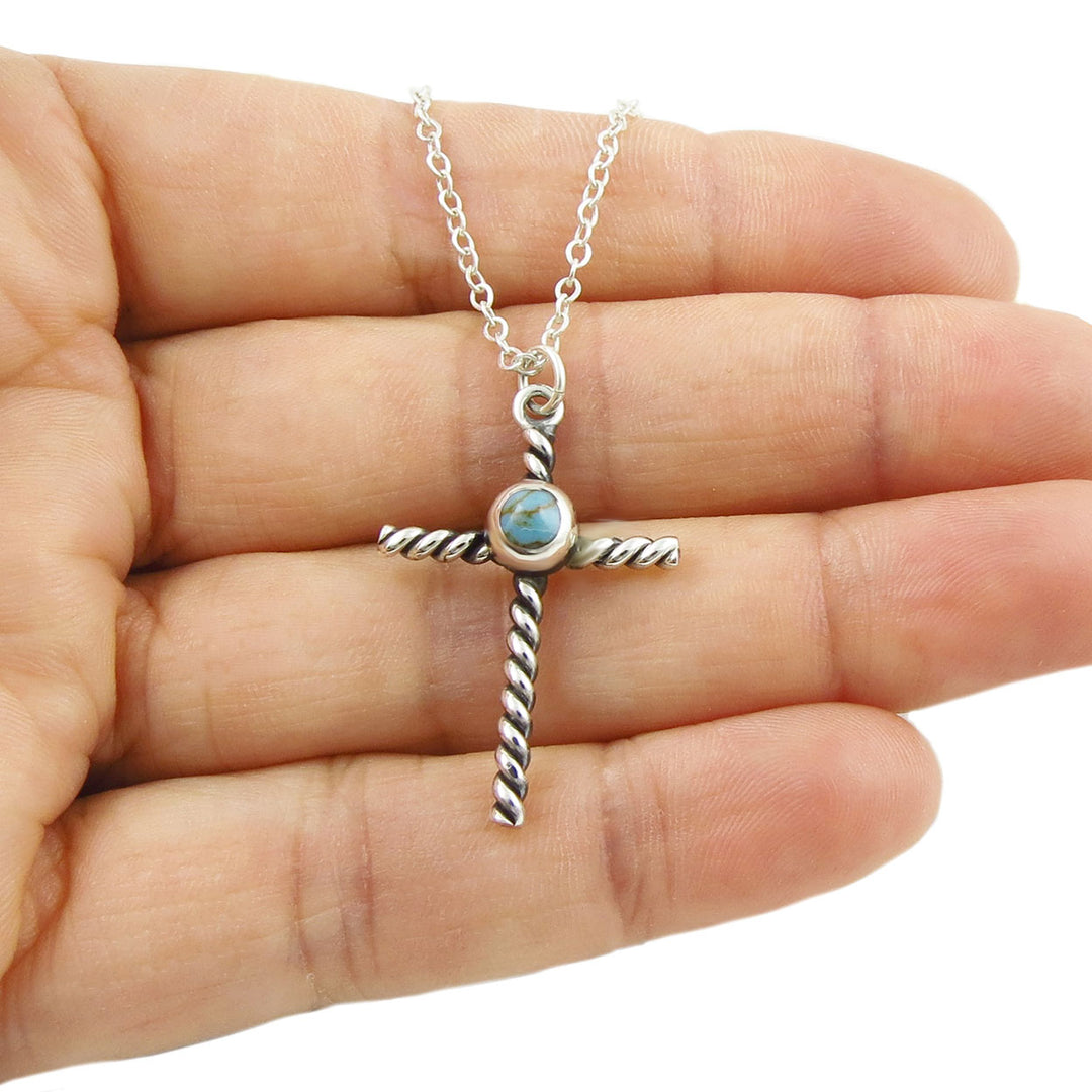 Silver cross pendant necklace with turquoise stone held in a hand on a white background
