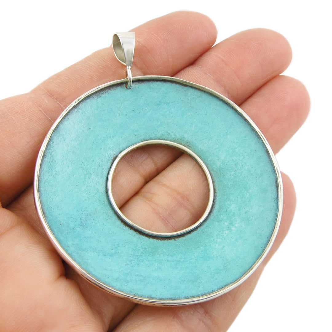 Large green copper patina pendant with silver frame held in a hand against a white background