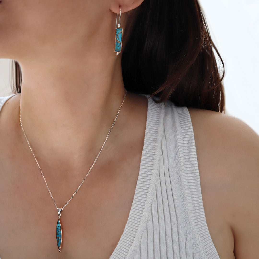 A woman in a white top with dark hair wearing a long chian necklace with a turquoise gemstone pendant and matching turquoise earrings, 