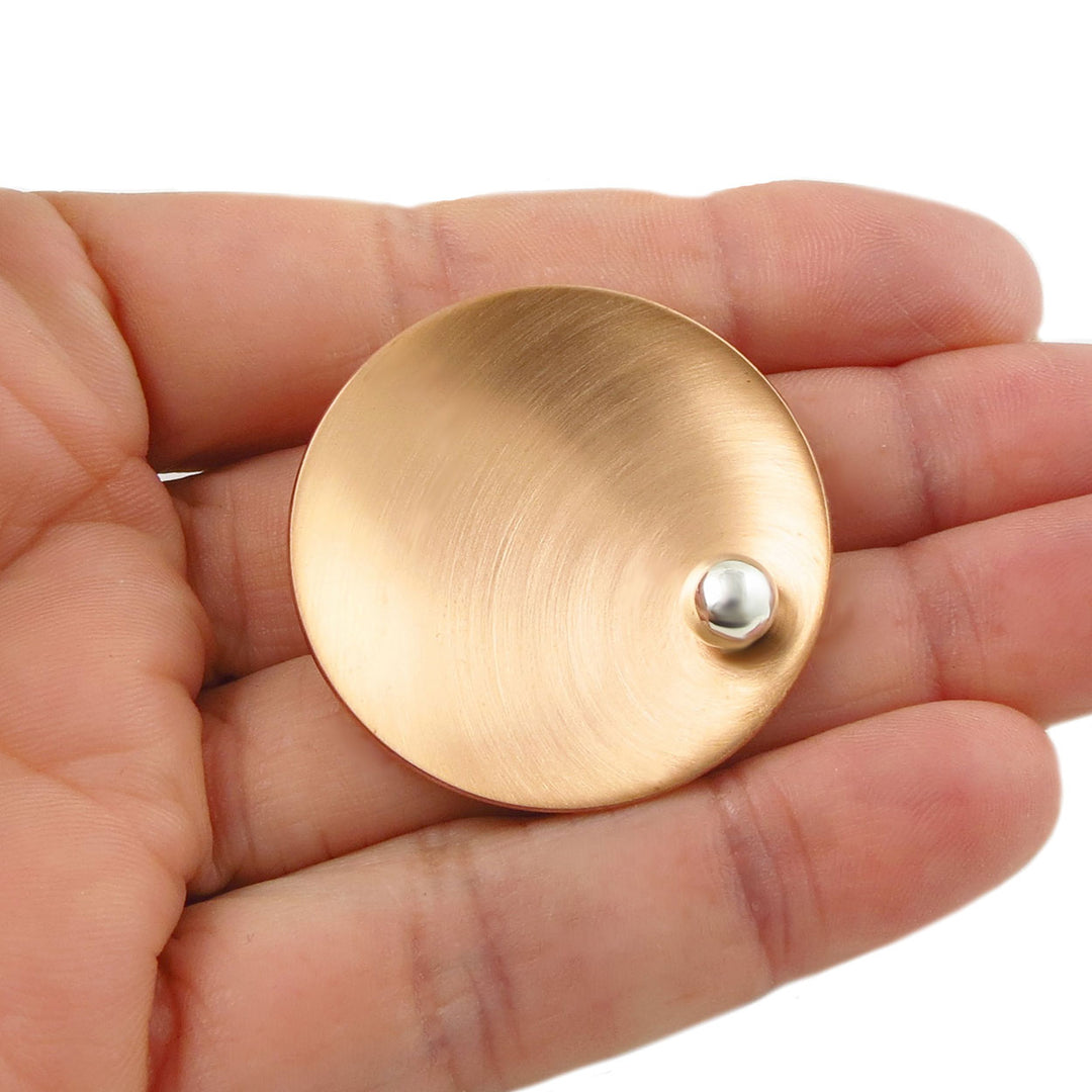 A large brushed copper pendant jewellery with a small silver ball accent held in a woman's hand on a white background