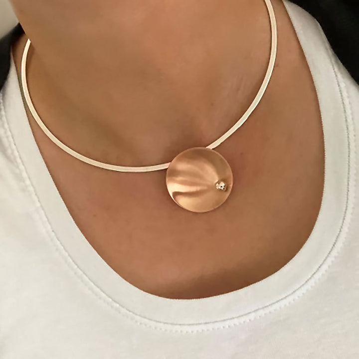 A brushed copper pendant and silver choker necklace worn by a woman in a white shirt.