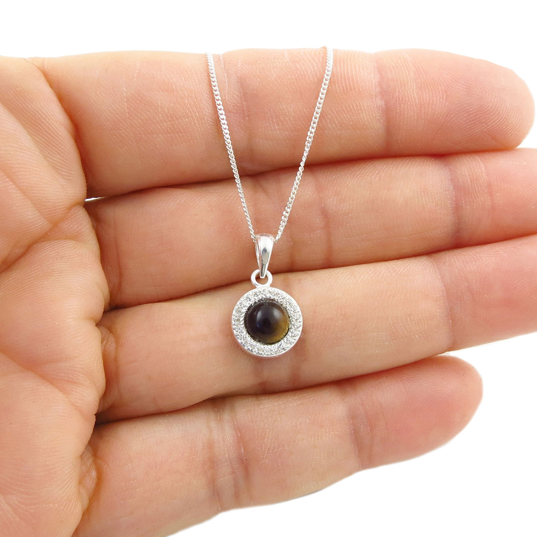 A sterling 925 silver pendant necklace with a round dark brown tiger's eye stone and a halo of small clear CZ stones, held in the palm of a hand against a white background.
