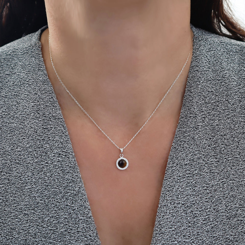 A close-up of a woman wearing a delicate 925 silver curb chain necklace with a small circular pendant featuring a dark tiger's eye gemstone and a halo of clear diamond-like stones, styled with a gray textured top. 