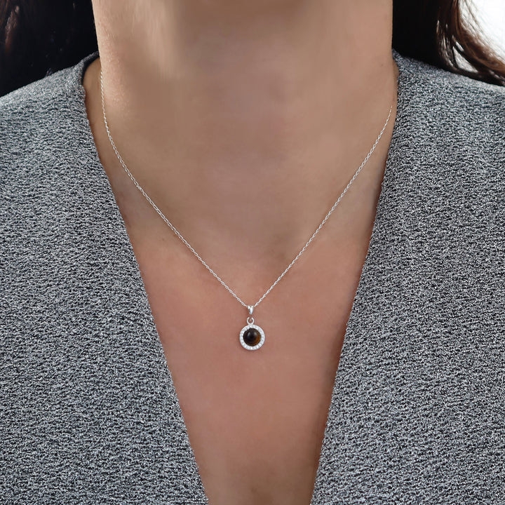 A close-up of a woman wearing a delicate 925 silver trace chain necklace with a small circular pendant featuring a dark tiger's eye gemstone and a halo of clear diamond-like stones, styled with a gray textured top. 
