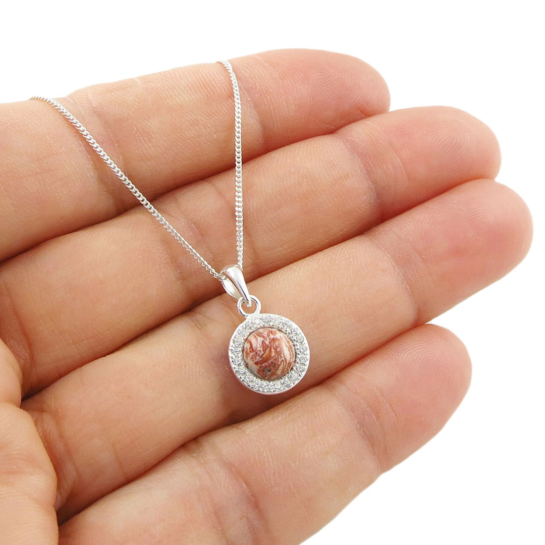 A sterling 925 silver chain necklace with a round pendant held in a hand. The pendant features a silver crystal halo with small pink rhyolite gemstone in the middle held in a person's hand. 