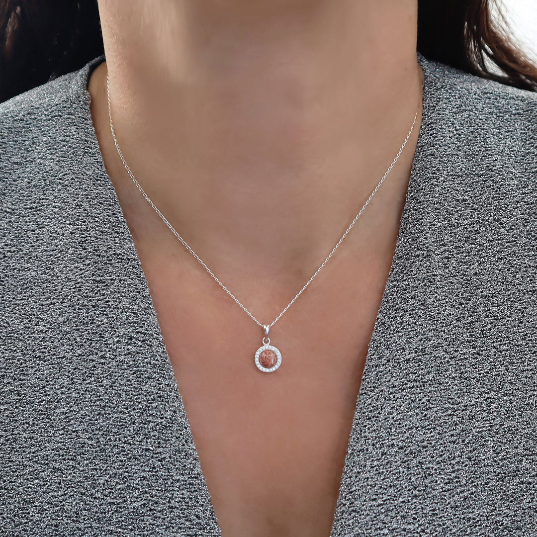 A close-up of a person wearing a delicate silver chain necklace with a small circular pendant featuring a pink rhyolite gemstone centre and a halo of clear stones, styled with a gray textured top.
